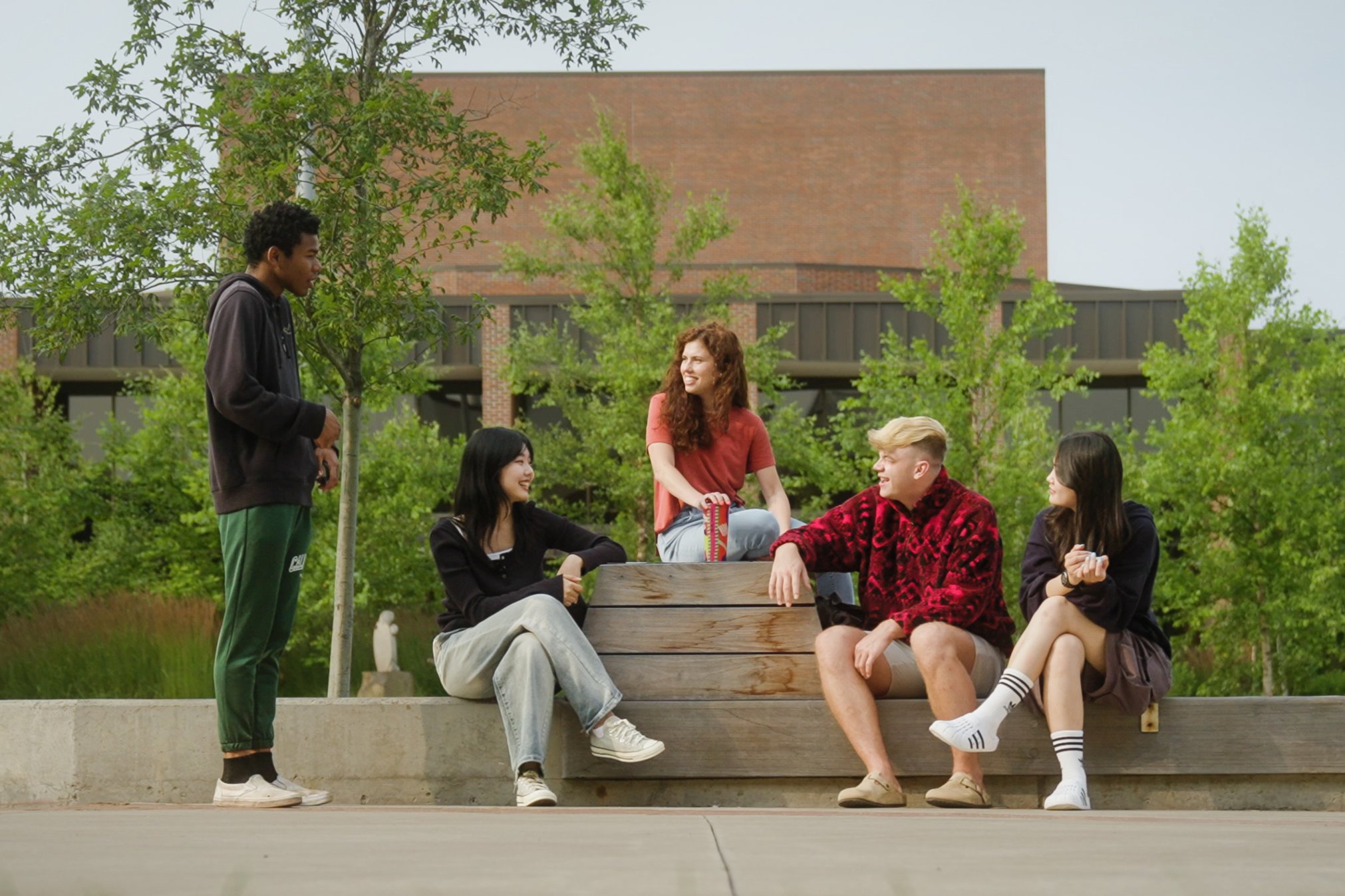 Five students laughing and talking together outside, with trees and campus buildings in the background. 