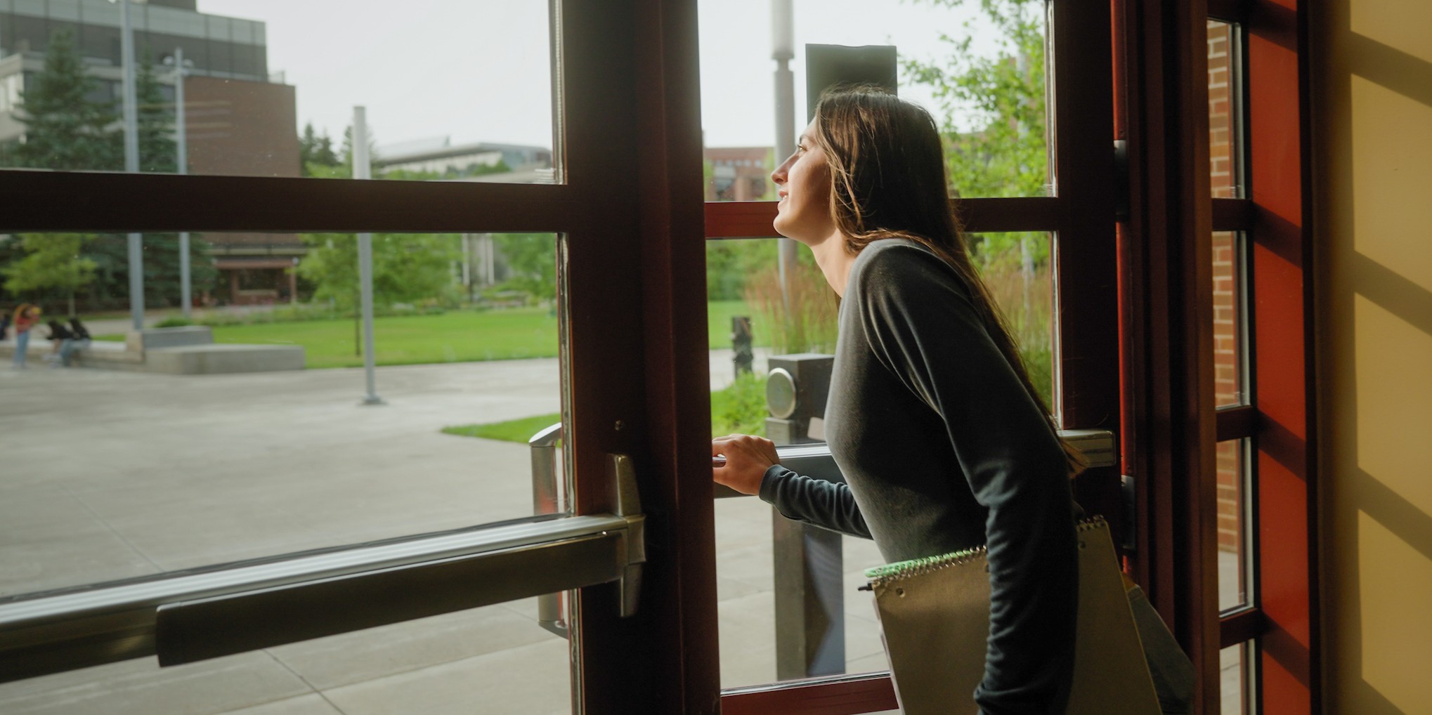 Sunlight shines on a student stepping through a doorway, with buildings and campus in the background.