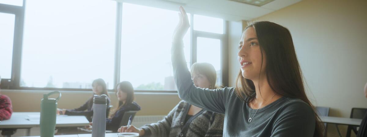 UMD student raises hand in classroom setting with bright, natural light coming through large window. 
